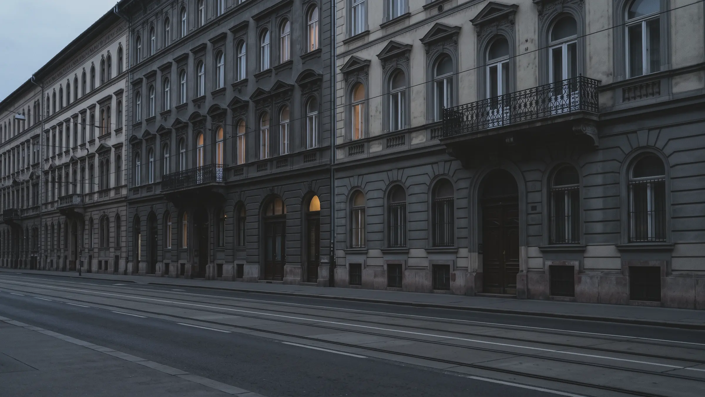 A street of tall stone apartment buildings has balconies, arched windows, and tram tracks, with lit windows at dusk.