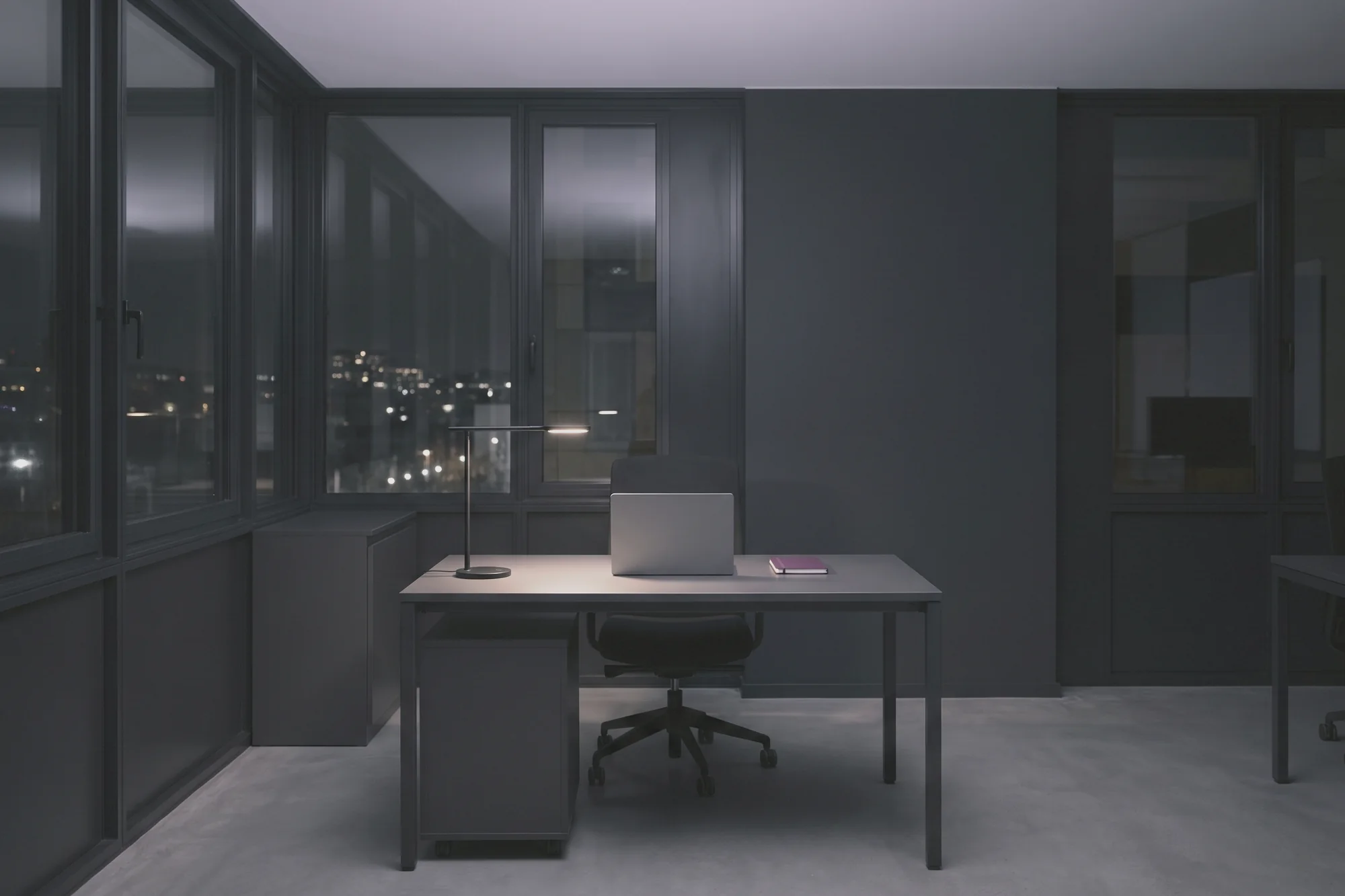 A single desk has an open laptop, a notebook and a small lamp, beside a glass wall overlooking city lights.