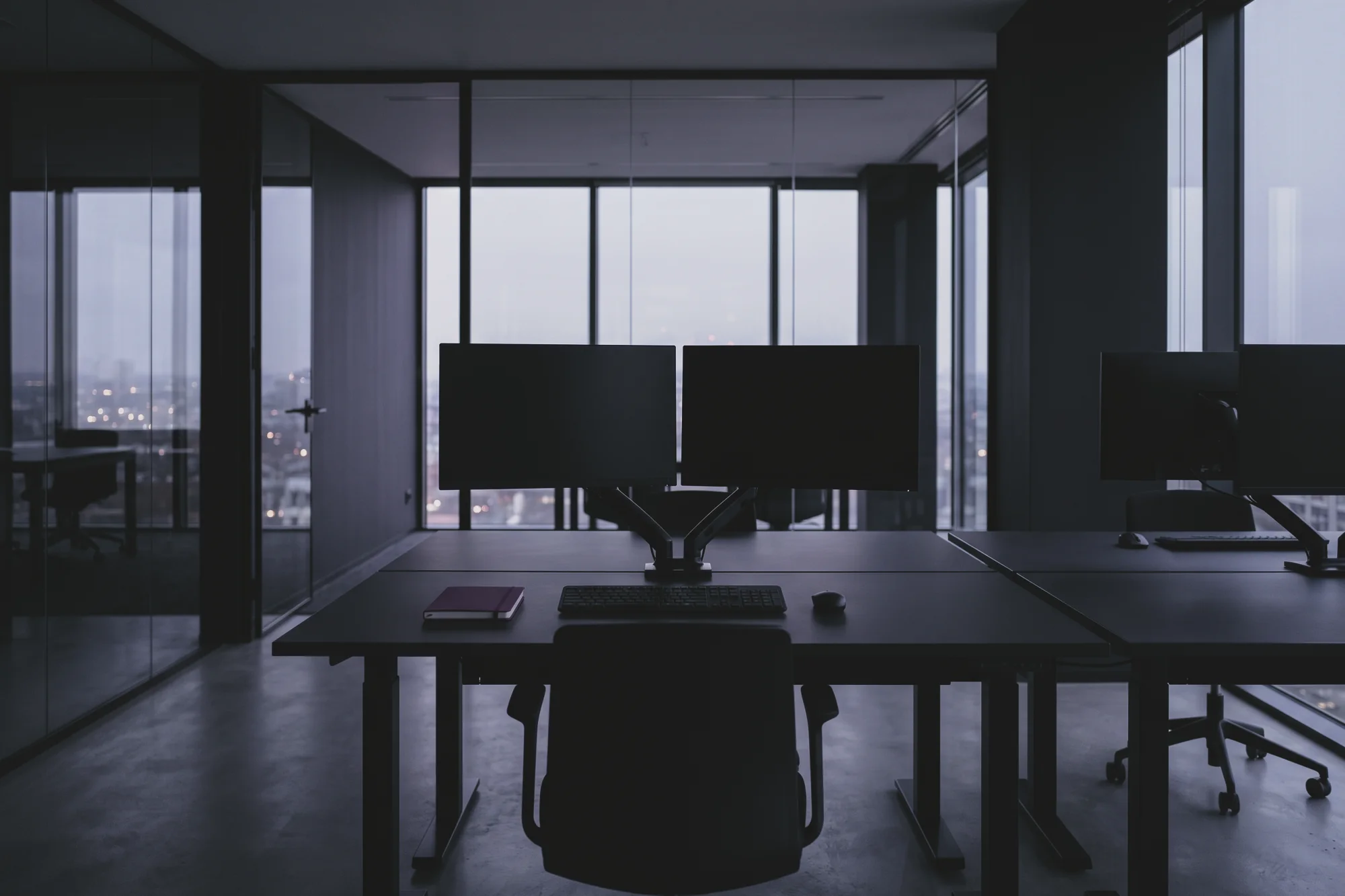 A desk has two monitors on arms and a chair by a glass wall, with a city view at dusk.