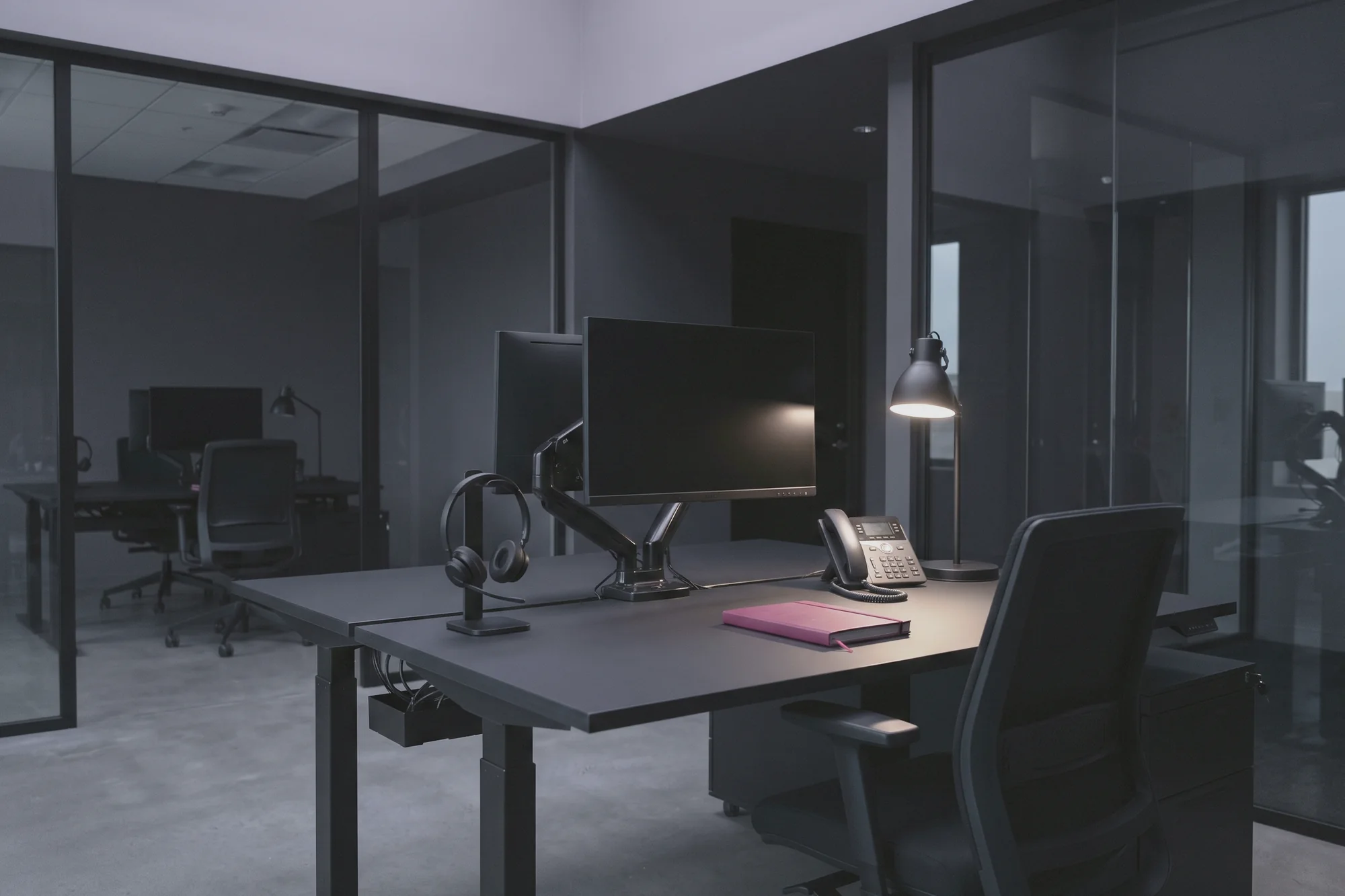 A desk has a monitor, a phone, a headset and a lamp, with more desks behind a glass partition.