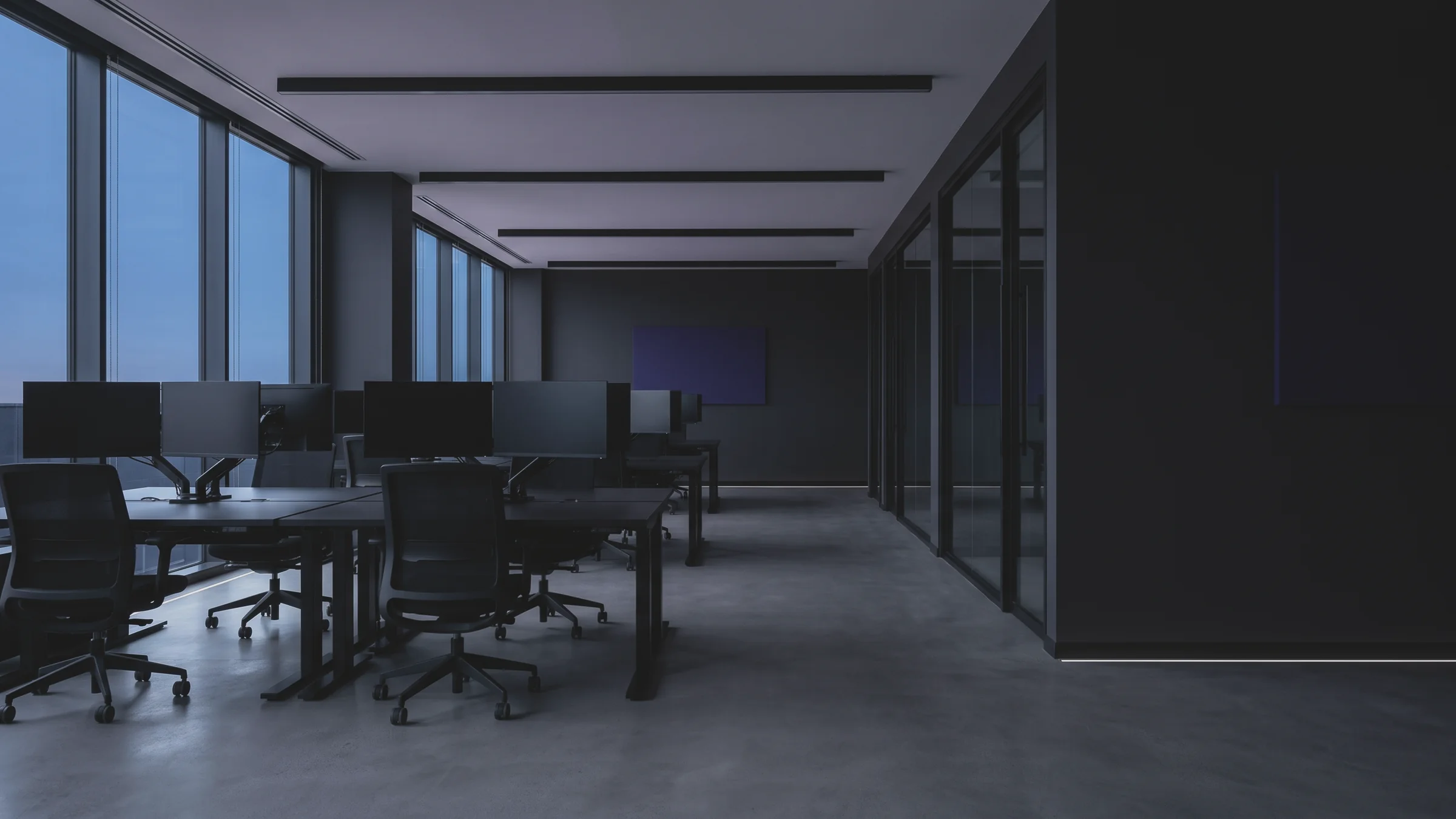 An open floor has desks with monitors and black chairs, beside tall windows showing the evening sky on the left.