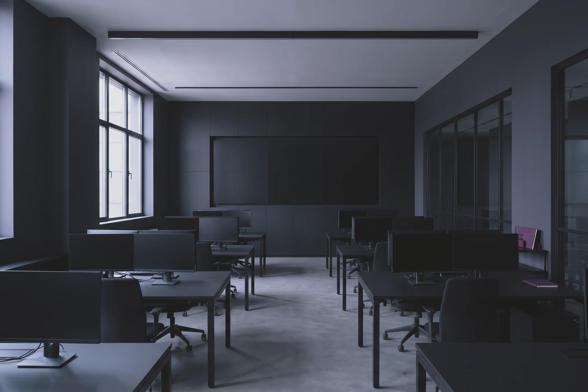 A large room of desks with monitors and chairs faces a dark wall display, beside tall windows on the left.