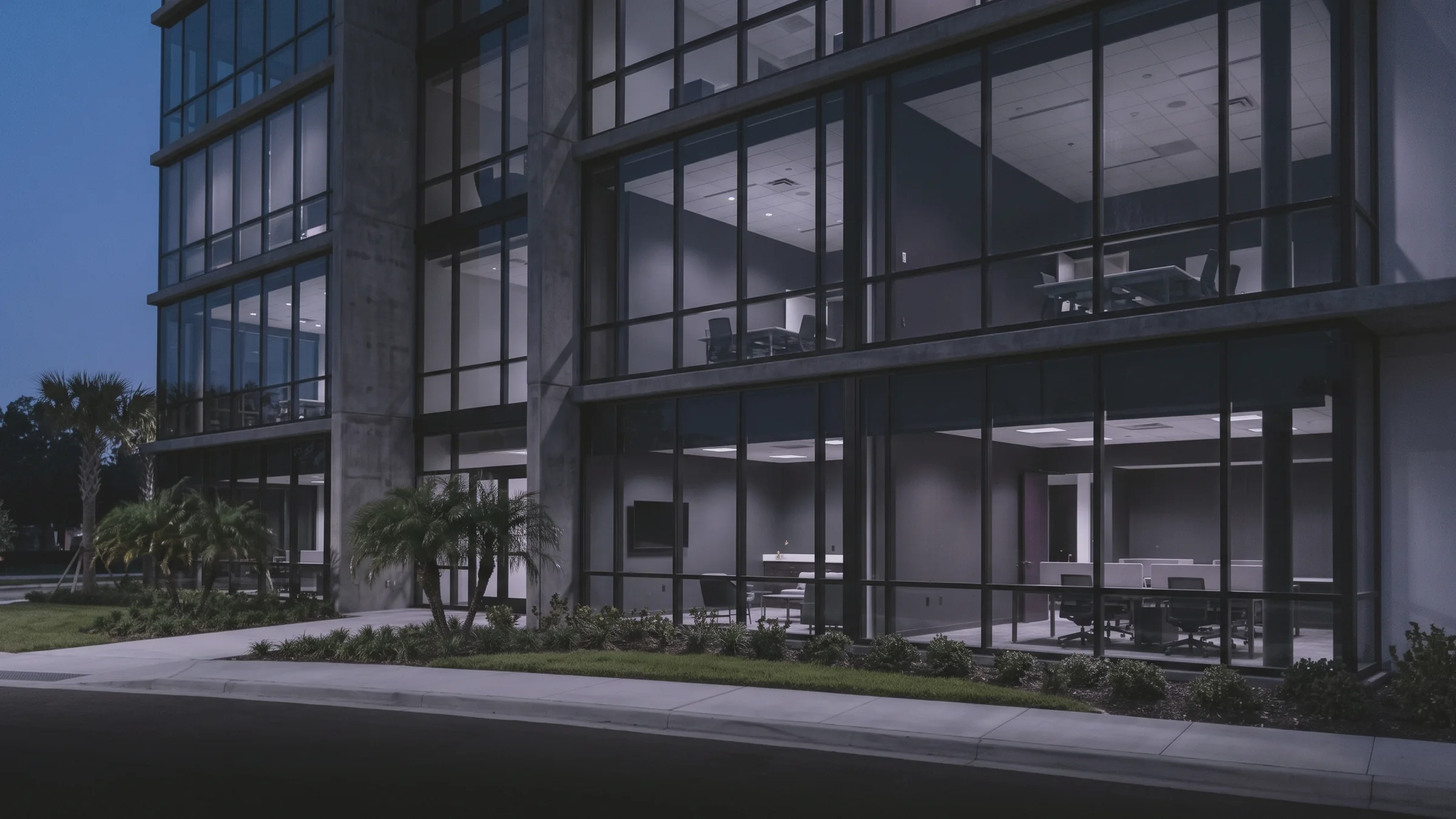 A modern glass and concrete office building stands at dusk, with palm trees, a lawn, and lit meeting rooms inside.
