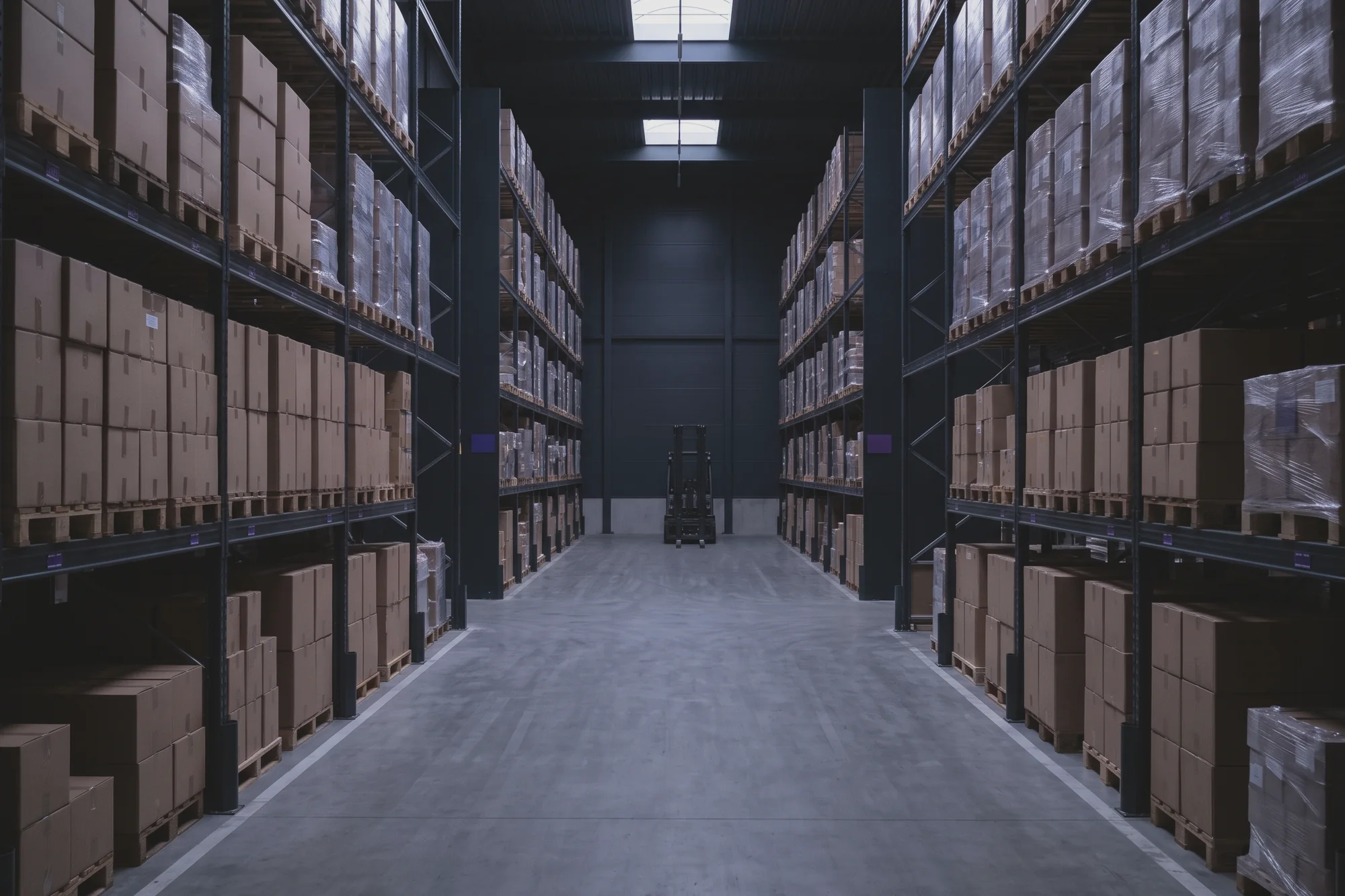 A wide warehouse aisle runs between tall racks of pallets stacked with boxes, and a forklift stands at the end.